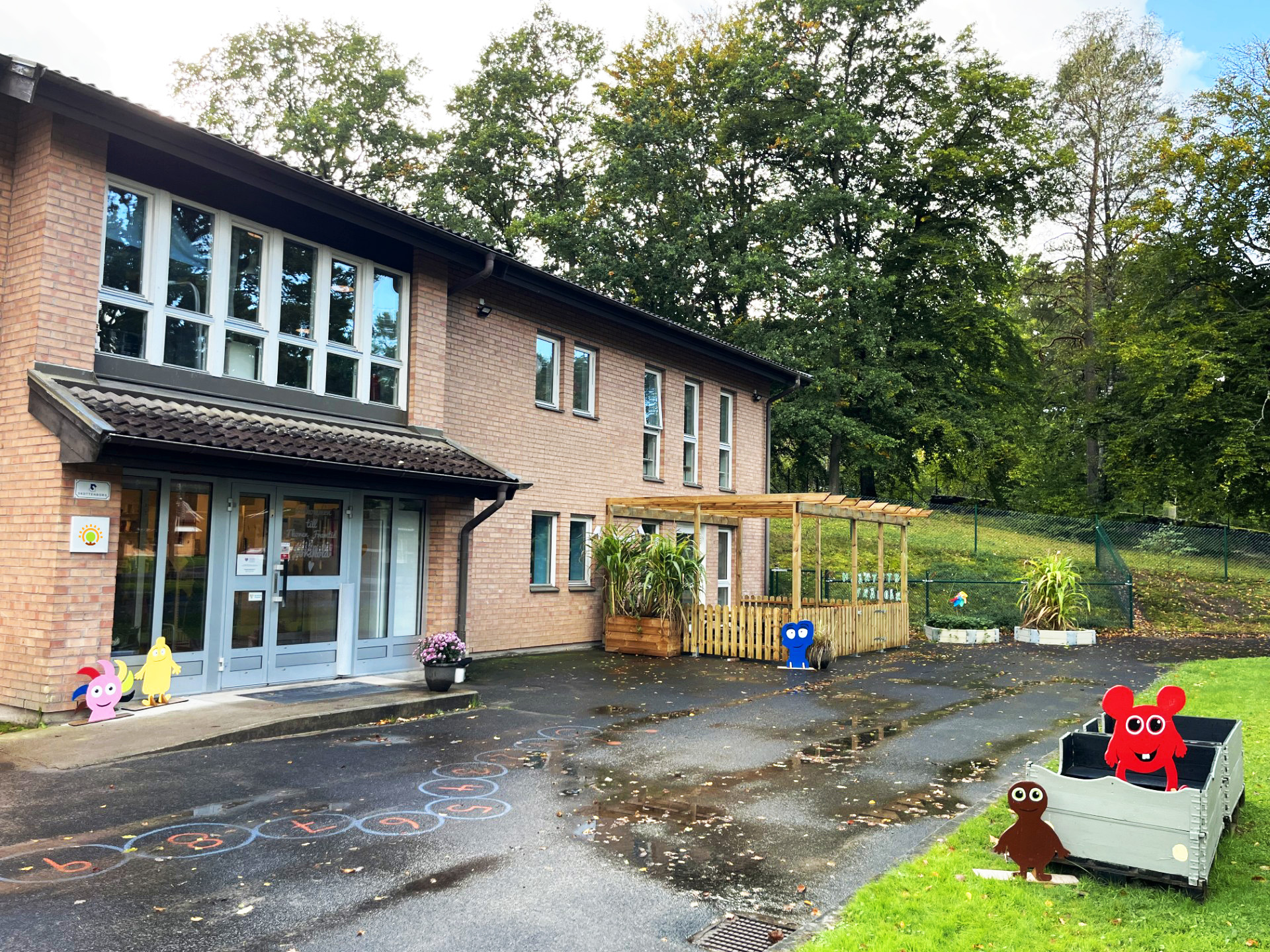 Fasad på en förskolebyggnad i tegel med grå entrédörrar och fönster i två våningar. På den asfalterade gården finns färgglada figurdekorationer i form av små monster, en hoppruta målad på marken, samt ett trädäck med staket och pergola. Höga lövträd omger byggnaden i bakgrunden.