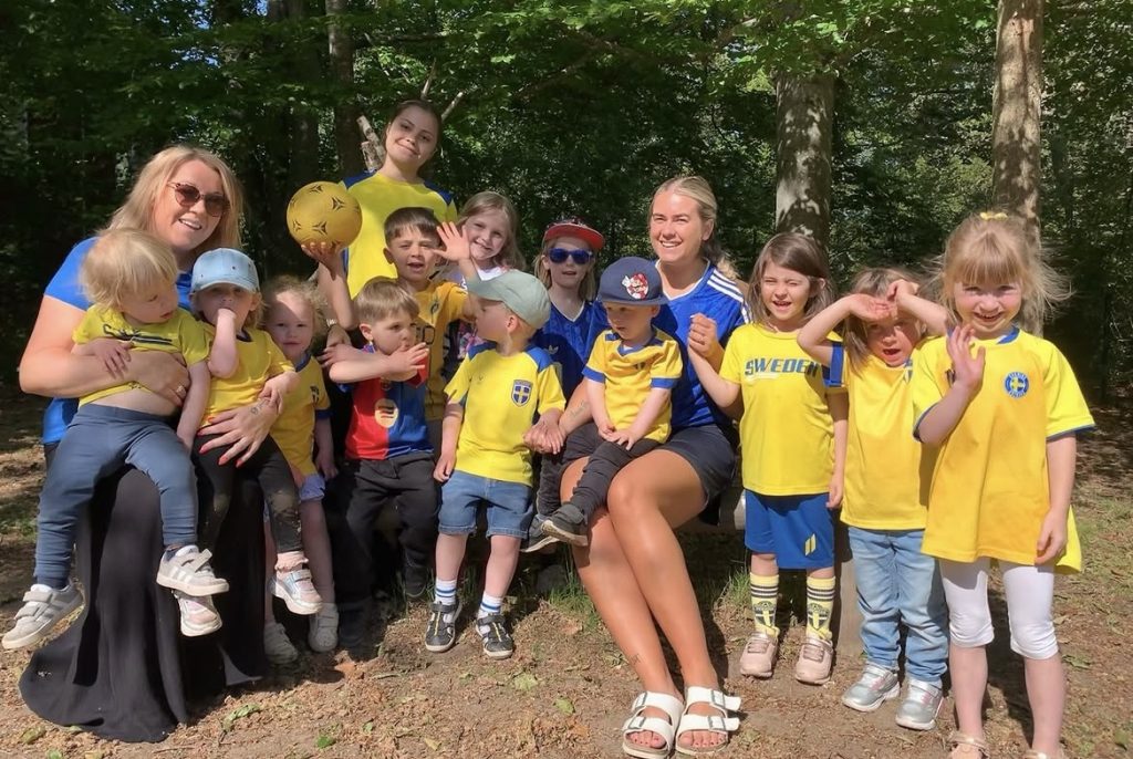 Grupp med barn och två vuxna utomhus i skogsmiljö, klädda i gula och blå fotbollströjor med Sverige-motiv. Flera barn vinkar och ler mot kameran, en person håller i en gul fotboll. Solljus filtreras genom trädkronorna i bakgrunden