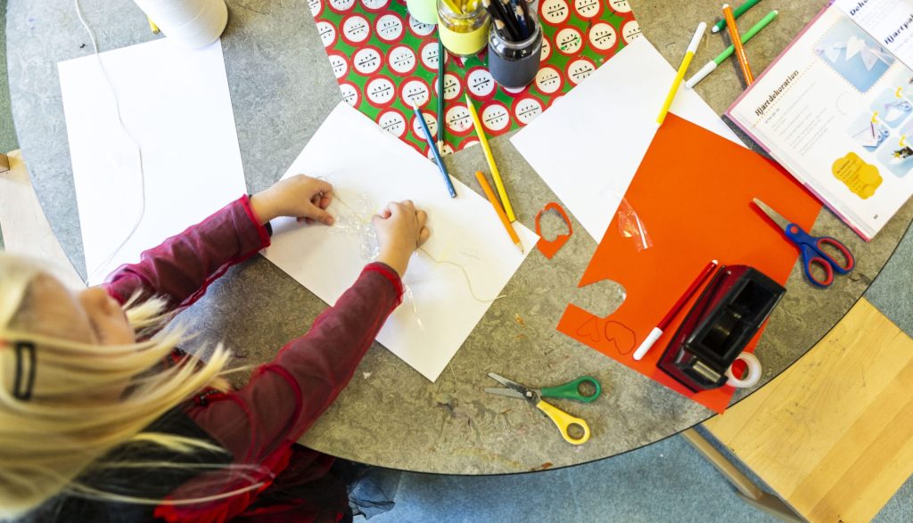 Vy uppifrån ett bord med pysselmaterial där ett barn med blont hår och röd tröja arbetar med papper och lim. På bordet ligger vita och orange papper, saxar, pennor, tejp och en instruktionsbok med texten 'Hjärtdekoration'
