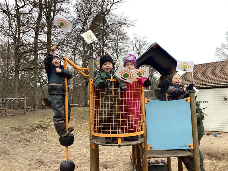 Barn sitter och ler inuti en koja byggd av pinnar och grenar i en kal höst- eller vinterskog. Ett tredje barn syns delvis gömt bland grenarna i bakgrunden. Marken är täckt av bruna löv.
