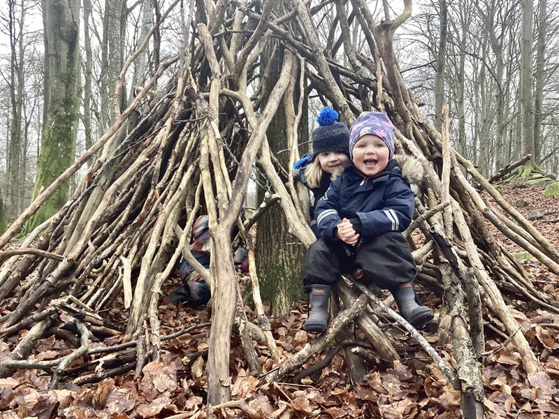 Barn sitter och ler inuti en koja byggd av pinnar och grenar i en kal höst- eller vinterskog. Ett tredje barn syns delvis gömt bland grenarna i bakgrunden. Marken är täckt av bruna löv.