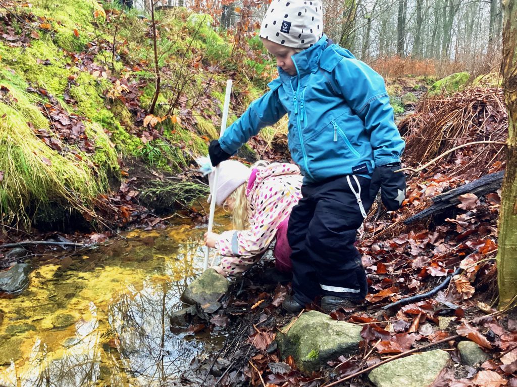 Två barn utforskar en bäck i skogen under hösten. Ett barn i blå jacka och mörka byxor står vid vattnet med en vit pinne, medan ett annat barn i ljus prickig jacka böjer sig ner mot vattnet. Marken är täckt av höstlöv och mossbeväxta stenar omger bäcken.
